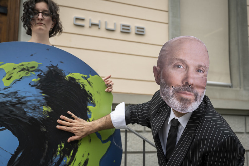 Someone wearing a mask of Chubb CEO Greenburg mask and a suit smears oil on a planet earth poster with CHUBB logo in the background
