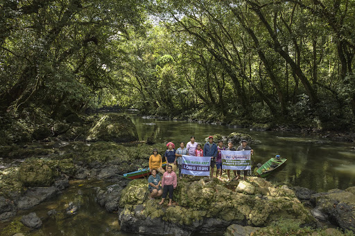 Long Isun community members standing together in the forest with banners that read "keep long isun forests standing" and "Akui Hutan Adat Long Isun"