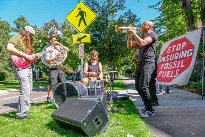 Brass band plays outside alongside the protest, in front of a banner that reads STOP INSURING FOSSIL FUELS
